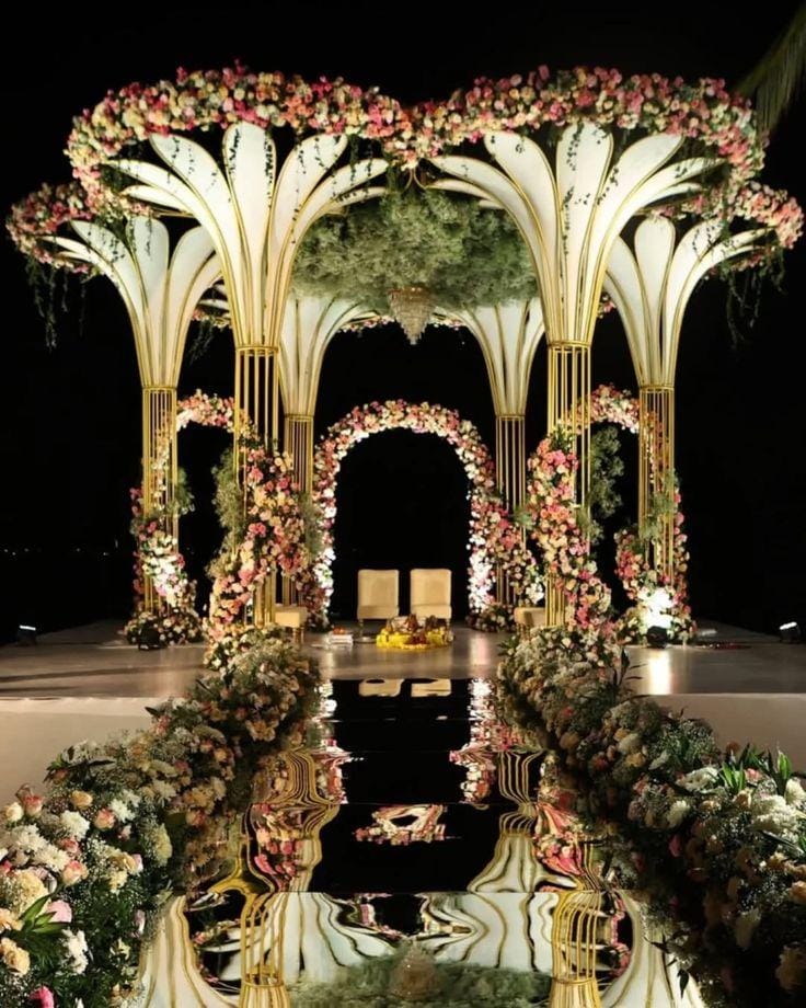 Reception stage with white palm-frond arches and a mirrored floral walkway by RAW Group